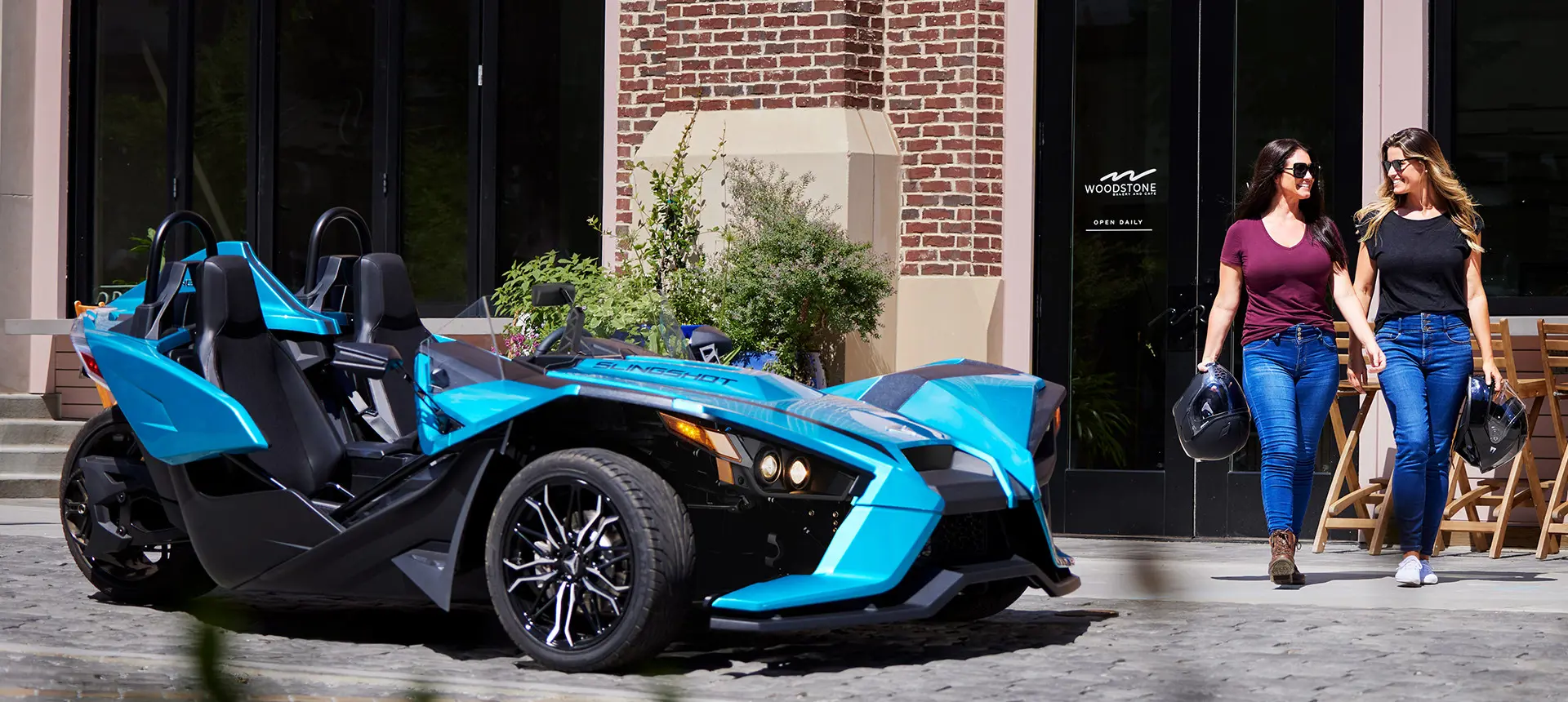 2 women walking in front of a pacific teal polaris slingshot sl three-wheeled motorcycle
