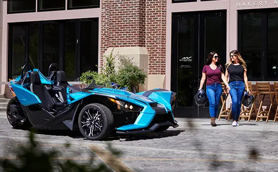 2 women walking in front of a pacific teal polaris slingshot sl three-wheeled motorcycle