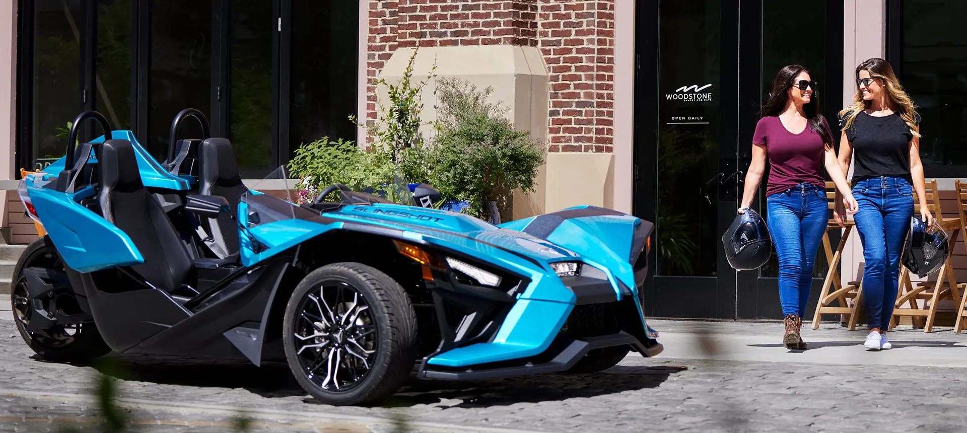 2 mujeres caminando frente a una motocicleta de tres ruedas polaris slingshot sl de color verde azulado pacífico