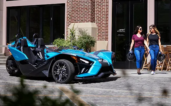 2 mujeres caminando frente a una motocicleta de tres ruedas polaris slingshot sl de color verde azulado pacífico