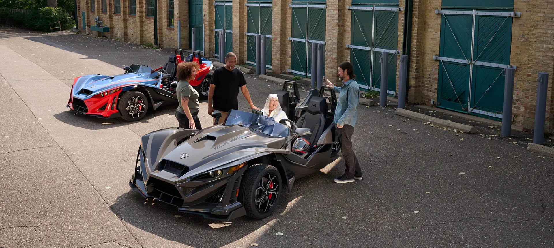 Aerial view of a woman sitting in the passenger seat of a 2025 Polaris Slingshot R three-wheeled roadster while 3 people stand near chatting.