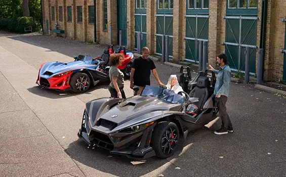 Vue du haut d’une femme assise sur le siège passager d’un cabriolet à 3 roues Polaris Slingshot R 2025 alors que trois personnes bavardent près d’elle.