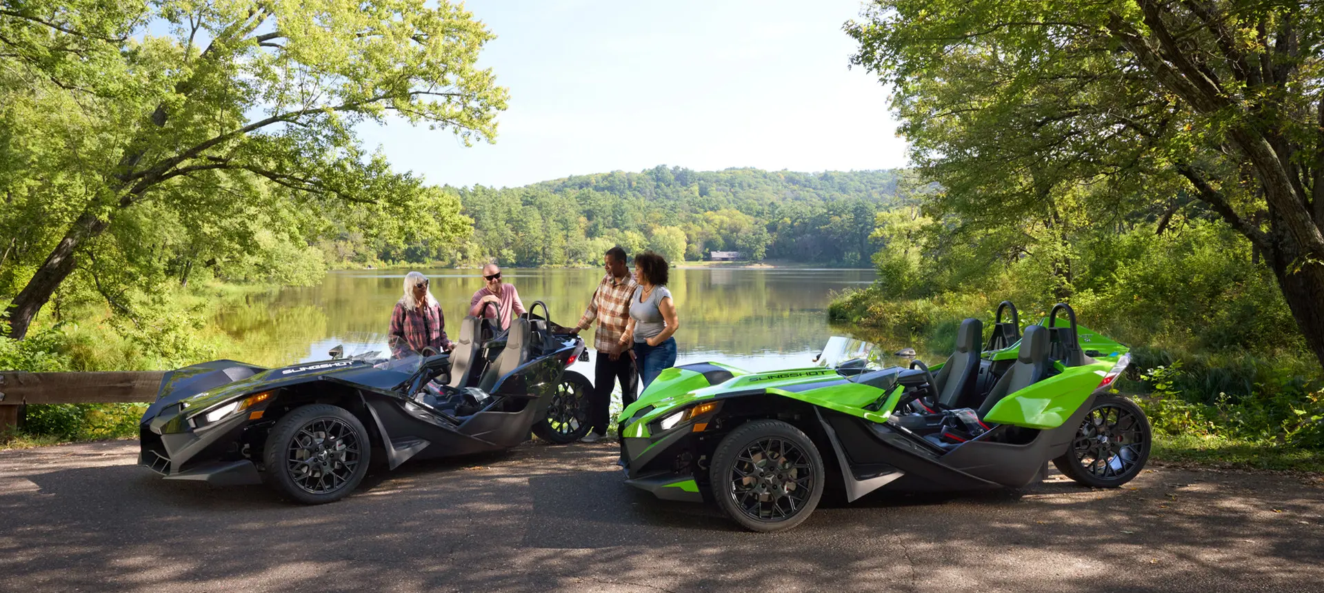 4 friends relaxing on shore by a lake near their 2025 Polaris Slingshot SL roadsters.