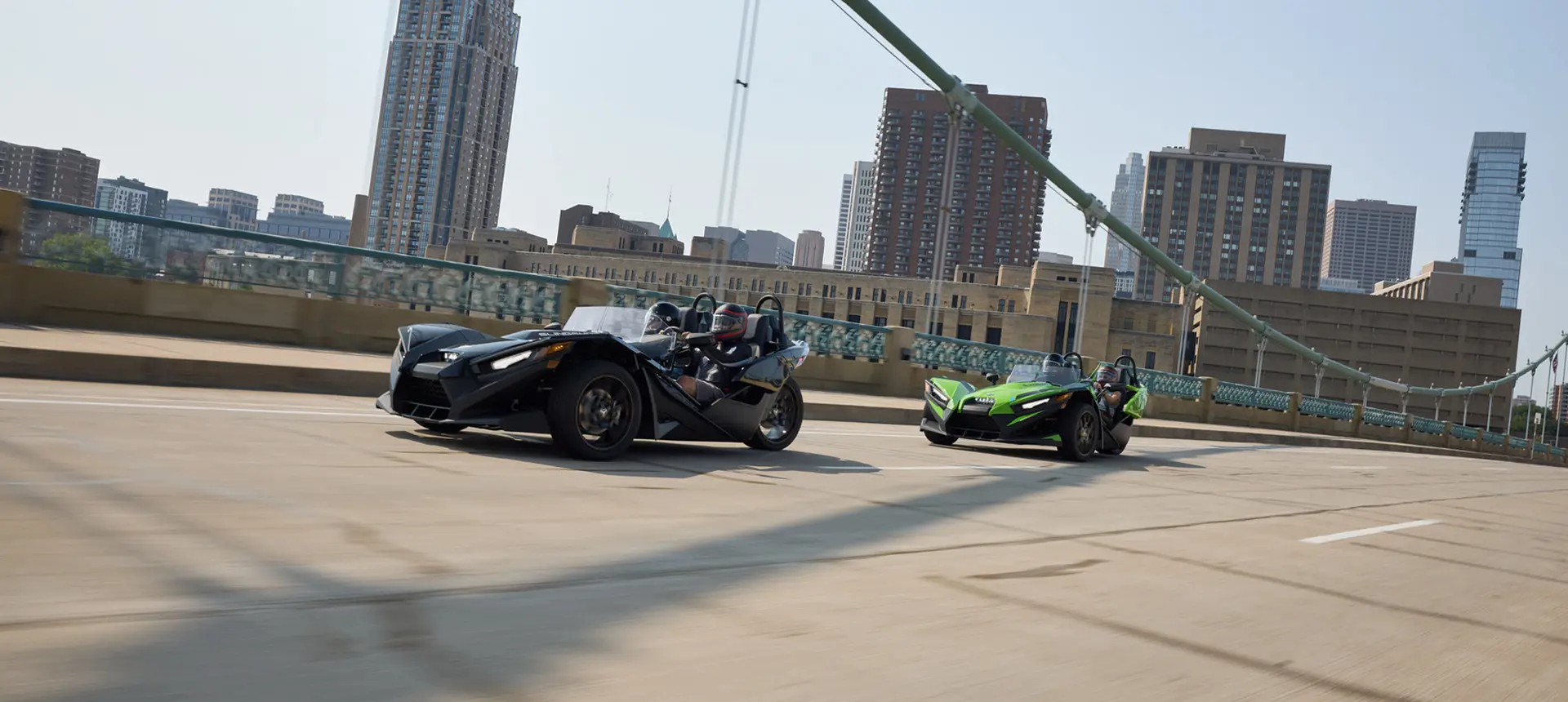 Two 2025 Polaris Slingshot SL three-wheeled motorcycles cruising across a bridge leaving a city behind.