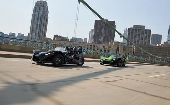 Two 2025 Polaris Slingshot SL three-wheeled motorcycles cruising across a bridge leaving a city behind.