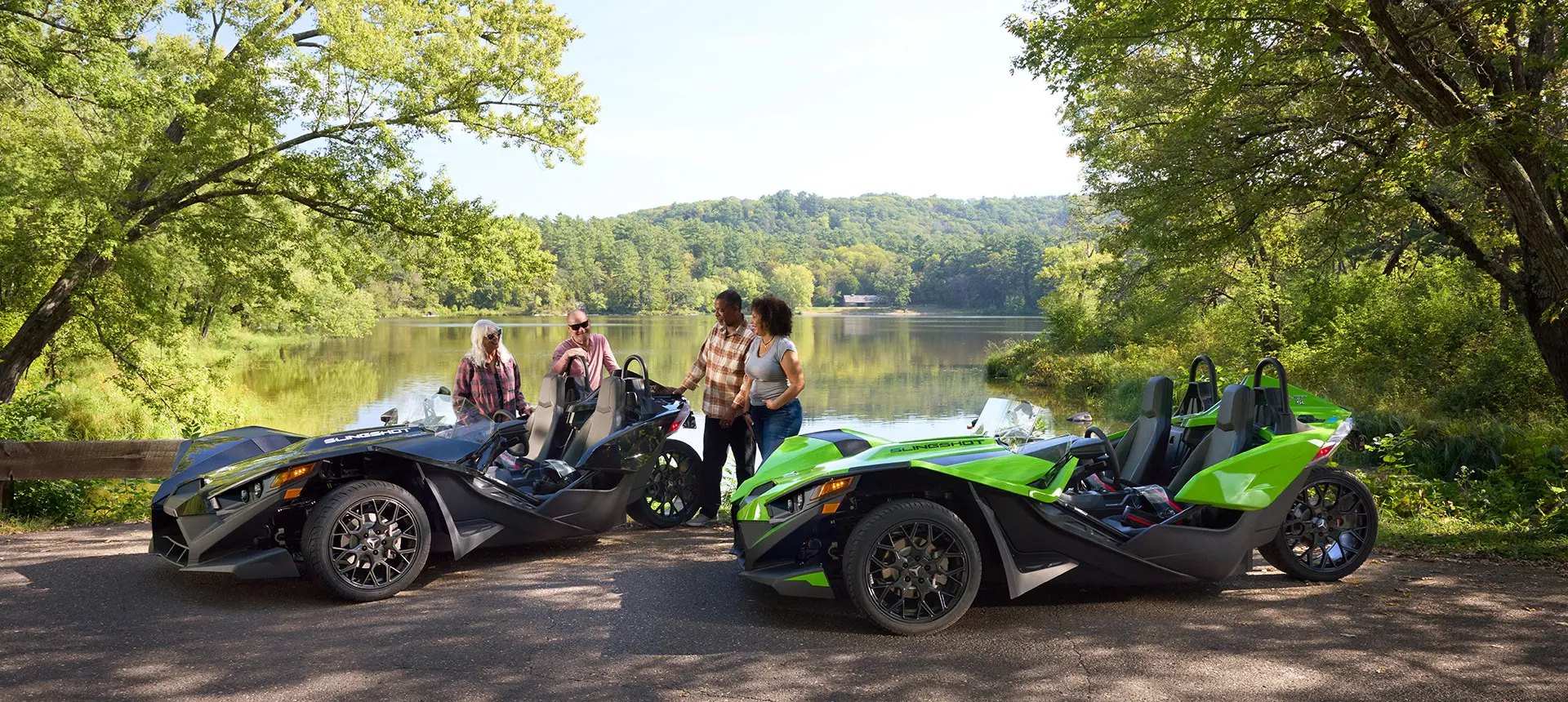4 friends relaxing on shore by a lake near their 2025 Polaris Slingshot SL roadsters.
