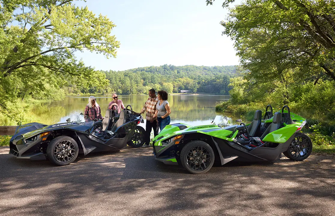 4 friends relaxing on shore by a lake near their 2025 Polaris Slingshot SL roadsters.