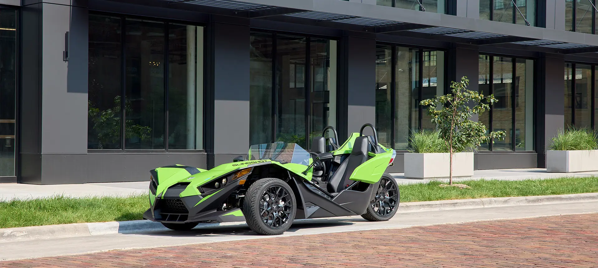 Profil latéral d’un cabriolet Polaris Slingshot SL 2025 Liquid Lime stationné à l’extérieur d’un bâtiment moderne.