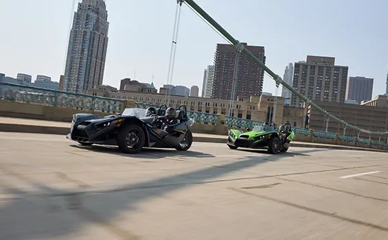 Two 2025 Polaris Slingshot SL three-wheeled motorcycles cruising across a bridge leaving a city behind.