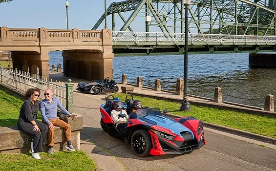 Two people with helmets on sitting in a 2025 Polaris Slingshot SLR three-wheeled motorcycle.