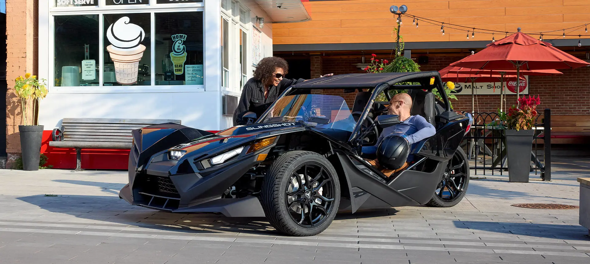 Hombre conversando con una mujer mientras está estacionado en un vehículo Polaris Slingshot de tres ruedas con mejoras de accesorios de las series Escape, Show, Drive y Design.