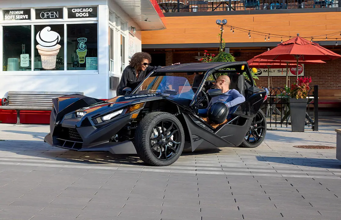 Man chats with woman while parked in a Polaris Slingshot three-wheeled vehicle with accessory upgrades from the escape, show, drive, and design series.