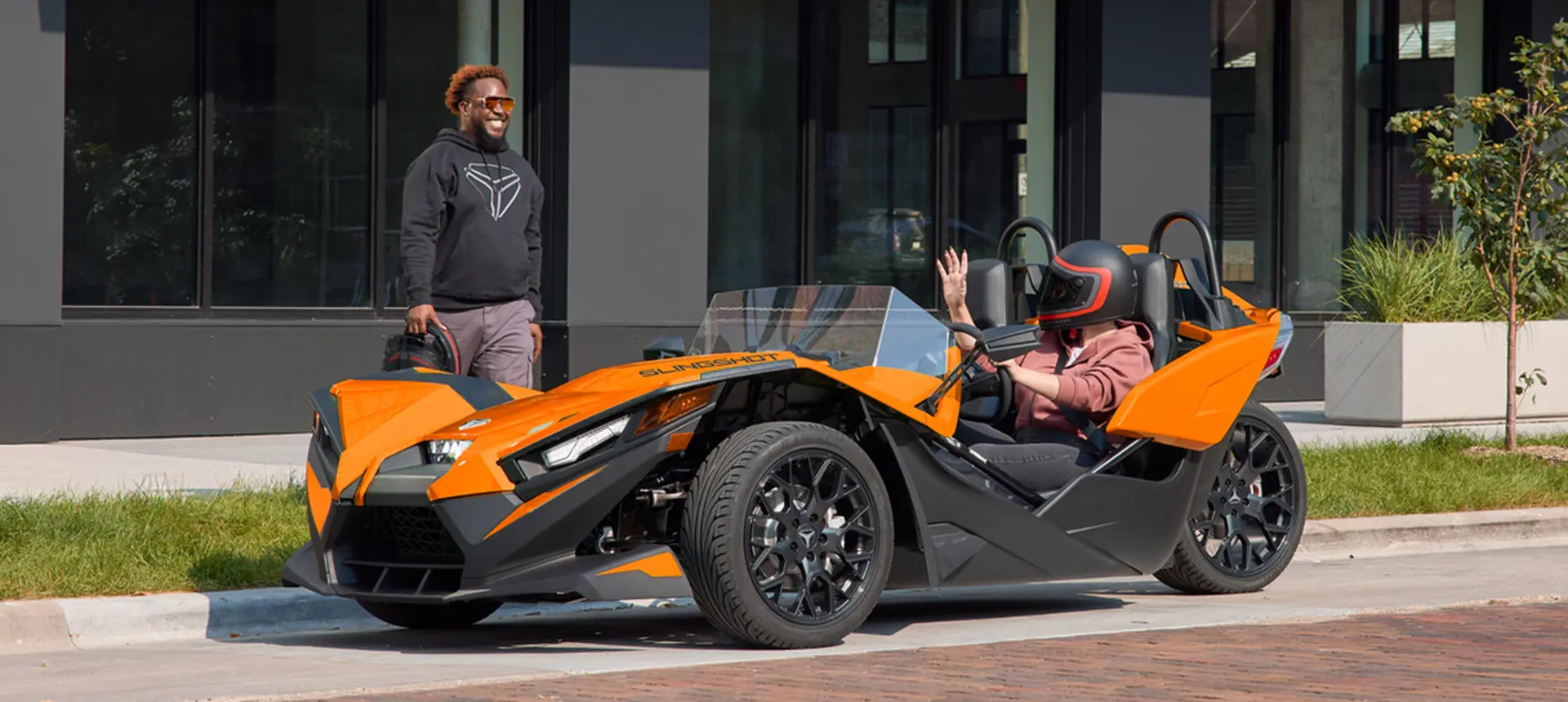 Hombre conversando con una mujer mientras está estacionado en un vehículo de tres ruedas Polaris Slingshot con mejoras de accesorios de las series Escape, Show, Drive y Design.