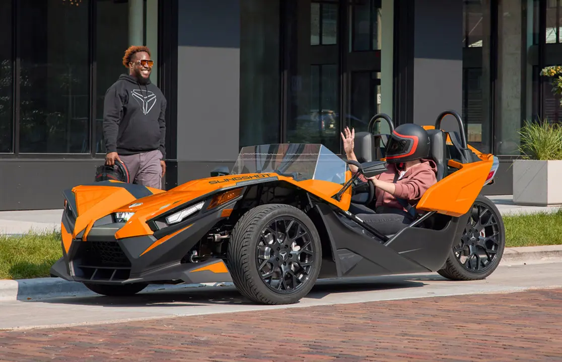 Hombre conversando con una mujer mientras está estacionado en un vehículo de tres ruedas Polaris Slingshot con mejoras de accesorios de las series Escape, Show, Drive y Design.