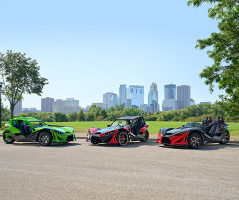 Three Polaris Slingshot vehicles in green, red/black, and black/red are parked on a paved road with a city skyline and green trees in the background under a clear sky.
