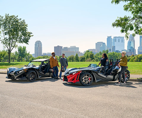 Three Polaris Slingshot vehicles in green, red/black, and black/red are parked on a paved road with a city skyline and green trees in the background under a clear sky.