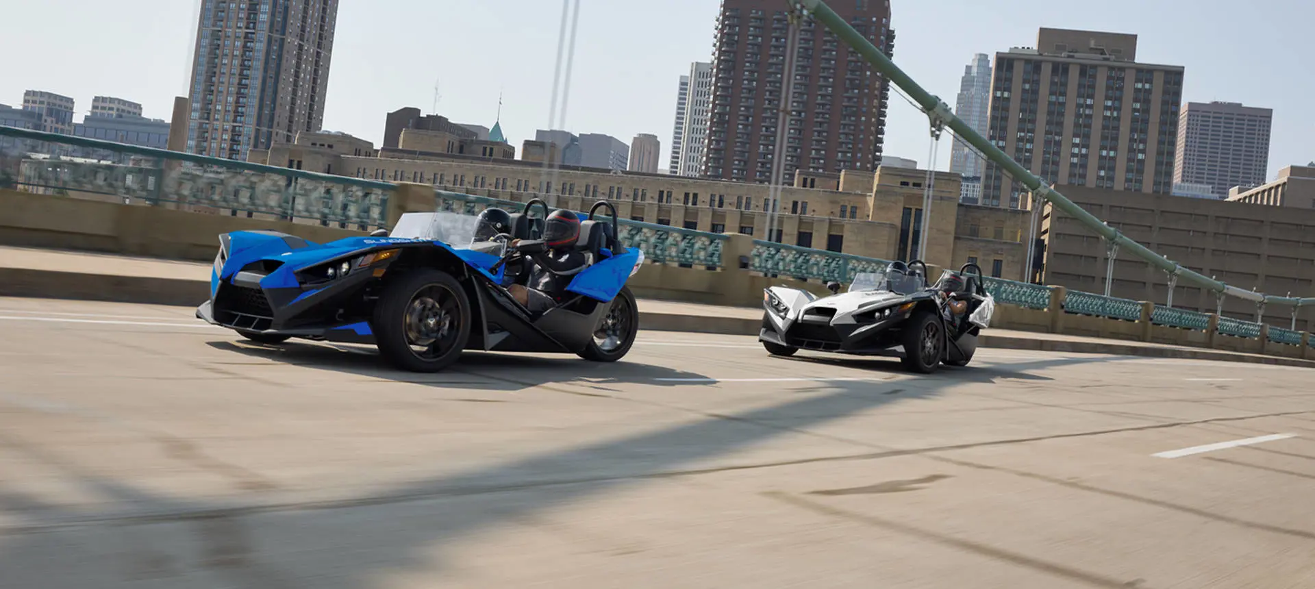Two 2026 Polaris Slingshot SL three-wheel roadsters cruising over a bridge near a city.