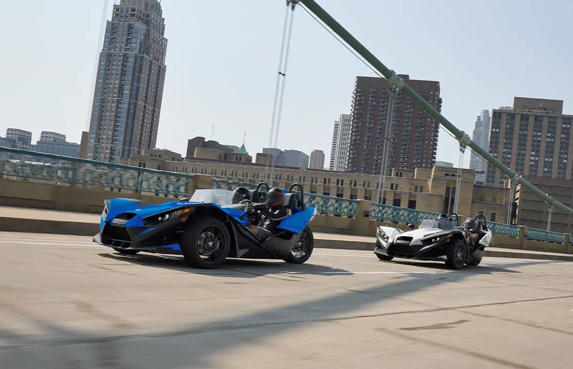 Two 2026 Polaris Slingshot SL three-wheel roadsters cruising over a bridge near a city.