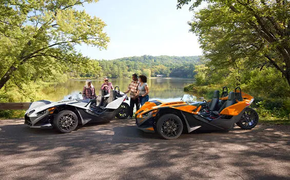 People standing near their parked 2026 Polaris Slingshot SL three-wheel motorcycles near a lake.