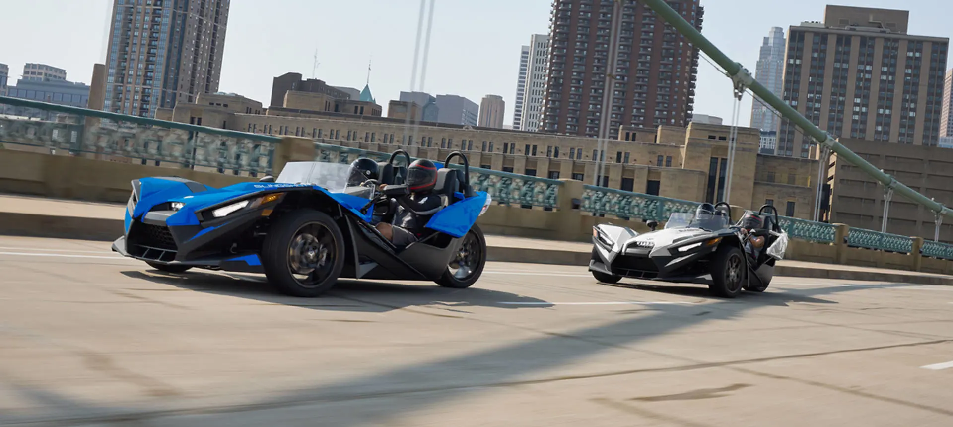 Two 2026 Polaris Slingshot SL three-wheel roadsters cruising over a bridge near a city.
