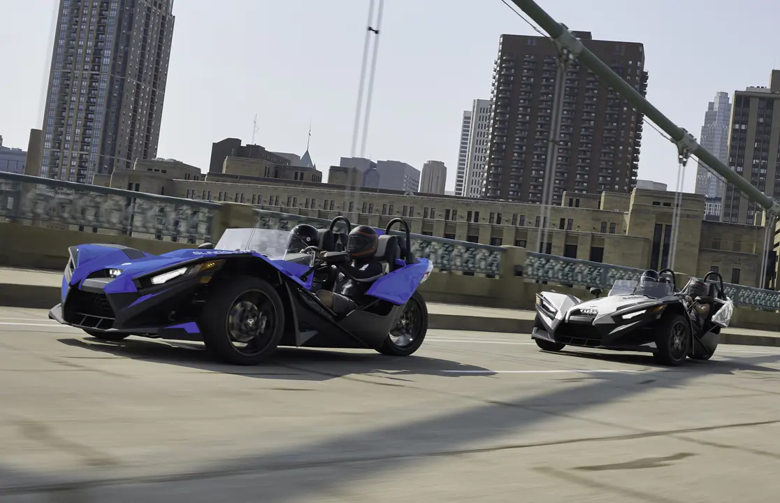 Two 2026 Polaris Slingshot SL three-wheel roadsters cruising over a bridge near a city.