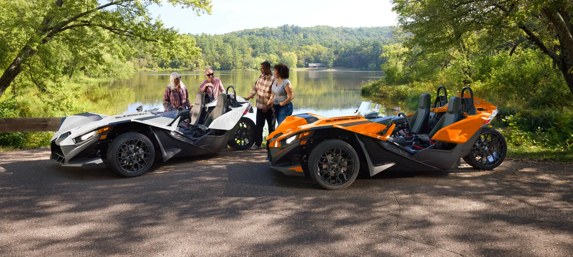People standing near their parked 2026 Polaris Slingshot SL three-wheel motorcycles near a lake.