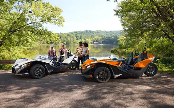People standing near their parked 2026 Polaris Slingshot SL three-wheel motorcycles near a lake.