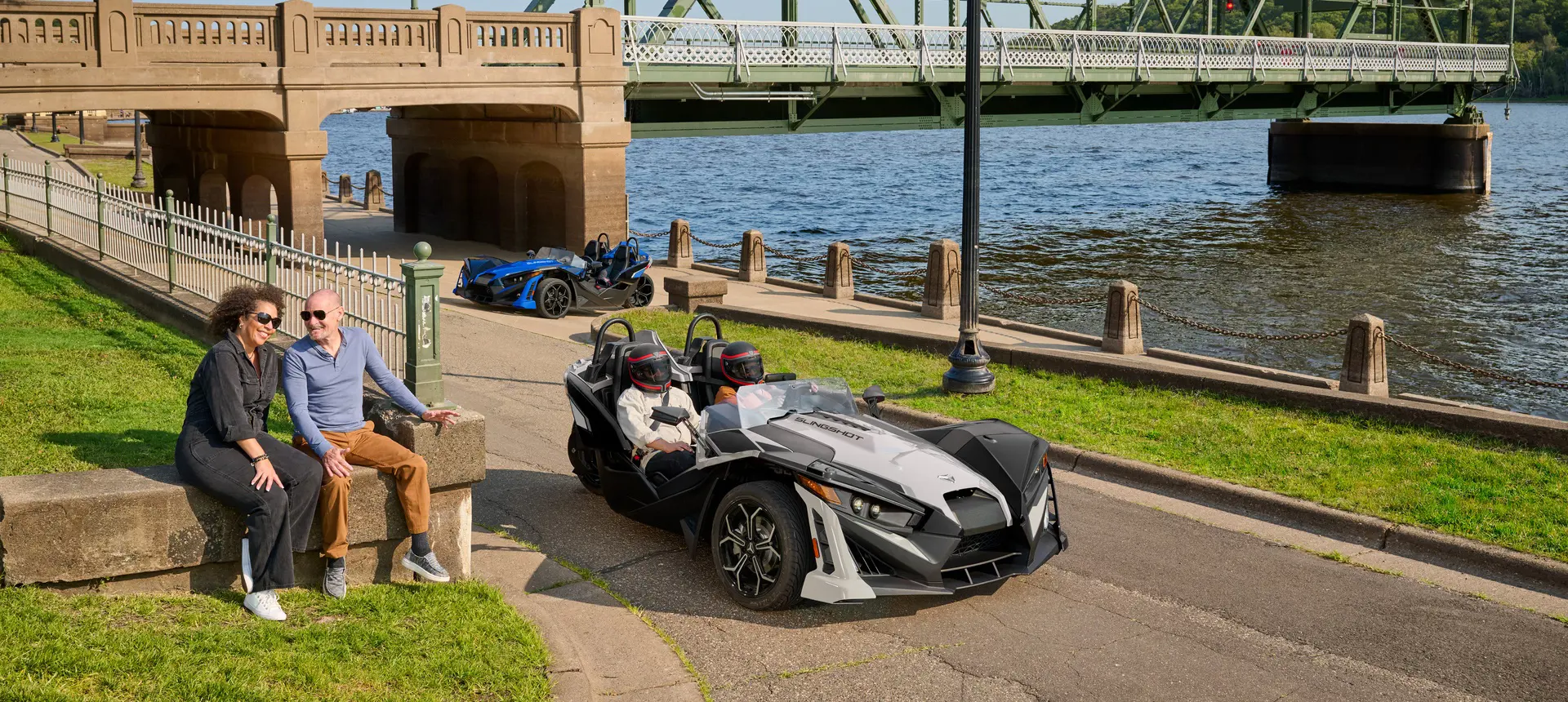 2 people driving a 2026 Polaris Slingshot SLR three-wheel roadster near a river.