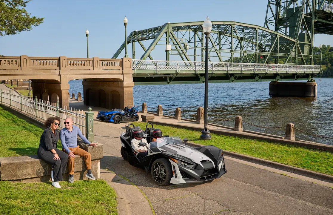 2 people driving a 2026 Polaris Slingshot SLR three-wheel roadster near a river.