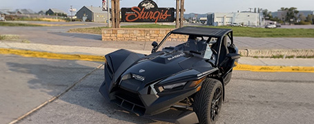 Black Polaris Slingshot parked on a street near a sign reading “Sturgis,” with buildings and open surroundings visible in the background.
