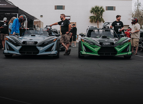 Polaris Slingshot vehicles displayed at an outdoor event, with multiple units parked side by side as attendees stand nearby and inspect the three-wheeled roadsters.