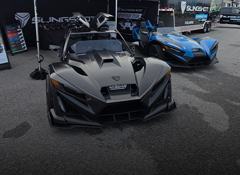 Two Polaris Slingshot vehicles on display at an outdoor event booth, one gray and one blue, parked on pavement beside signage and branded trailers.
