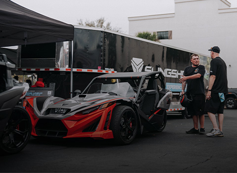 Polaris Slingshot vehicle displayed at an demo event, with two attendees standing beside it near a branded trailer and discussing features.