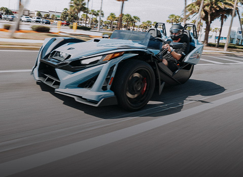 Polaris Slingshot driving on a city street, captured in motion with a helmeted driver, palm trees, and urban buildings blurred in the background.