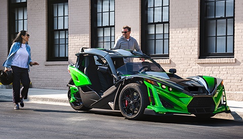 Two modern Polaris Slingshot three-wheeled vehicles racing on a sunny road, surrounded by lush greenery.