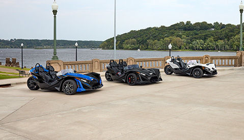 A red and black Polaris Slingshot three-wheeled roadster parked on a concrete rooftop, with a modern urban cityscape and tall buildings in the background.