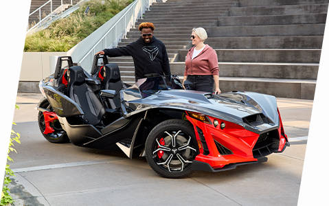 A red and black Polaris Slingshot three-wheeled vehicle with two occupants, parked outdoors near stairs, with the driver standing beside it.