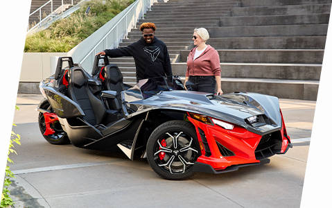 Un vehículo de tres ruedas Polaris Slingshot rojo y negro con dos estacionados al aire libre cerca de unas escaleras, con el conductor y el pasajero parados junto a él. 