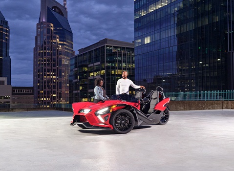 Two people standing around a red and black Slingshot. 