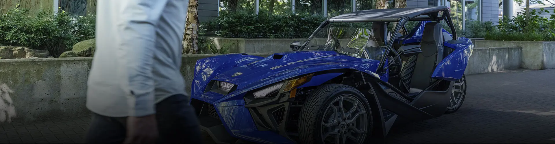 Blue Polaris Slingshot three-wheel roadster parked outside a modern building as a person walks past in the foreground.