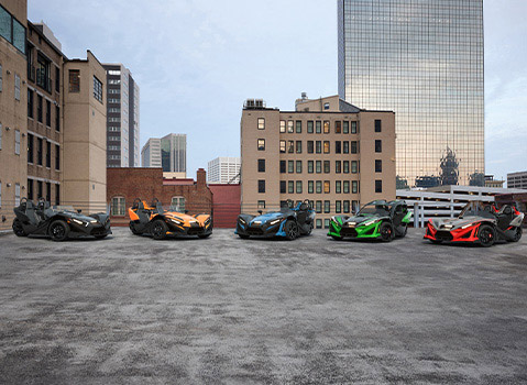 Five Polaris Slingshot three-wheel roadsters in black, orange, blue, green and red displayed on a rooftop against a city skyline.