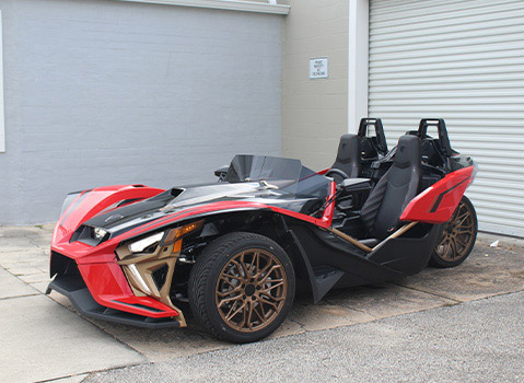 Customized red and black Polaris Slingshot three-wheel vehicle with bronze wheels parked outside a garage.