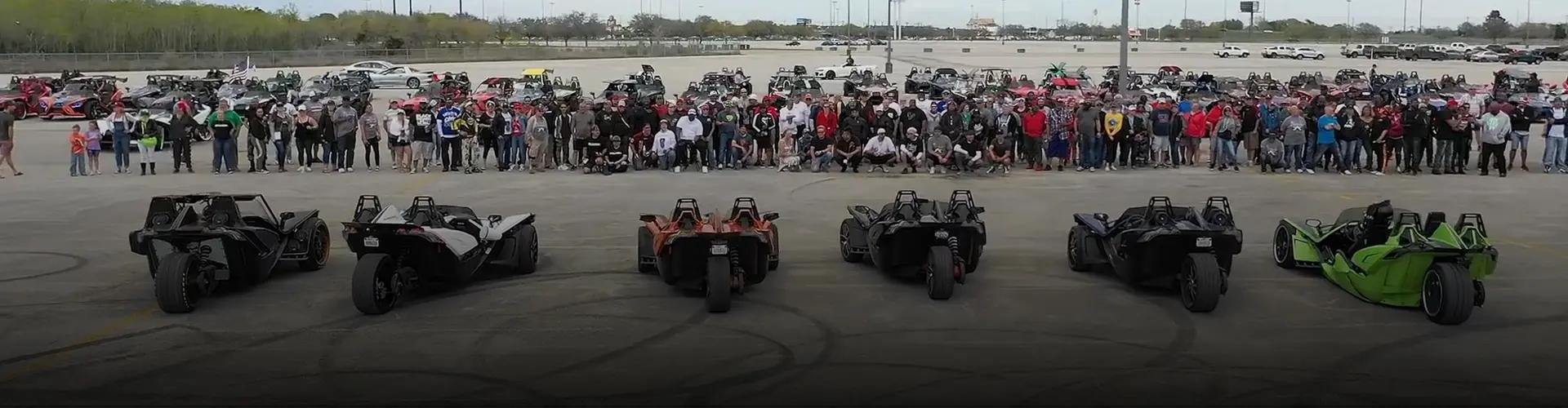 Large group gathered behind six Polaris Slingshot autocycles at an outdoor owners’ event in a parking lot.