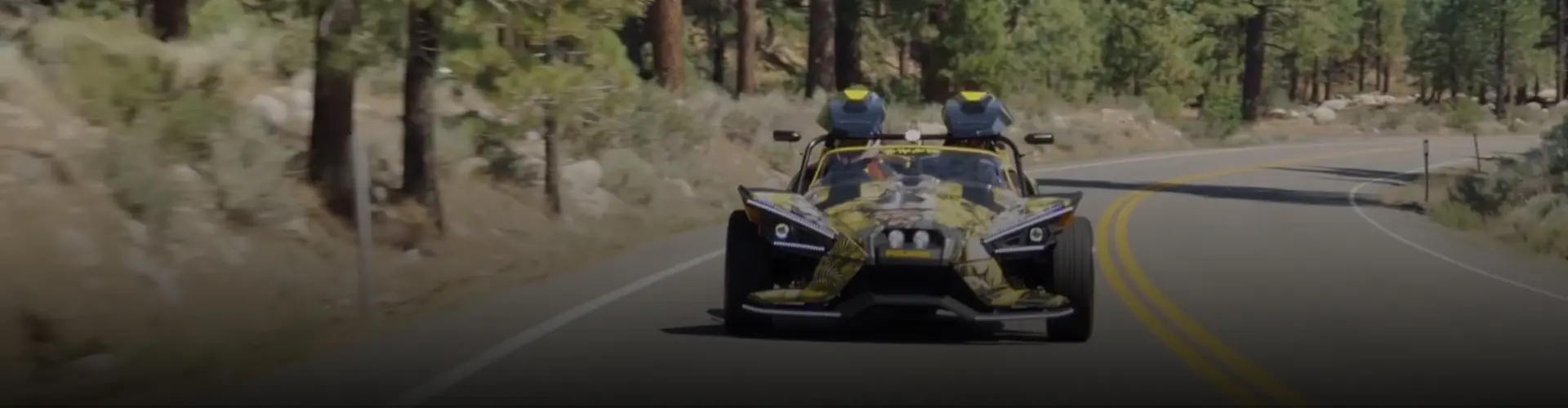 Custom Polaris Slingshot three-wheel vehicle driving along a winding road through a pine forest.