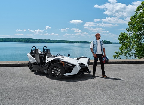 Man holding a helmet beside a white Polaris Slingshot roadster parked at a scenic lakeside overlook.