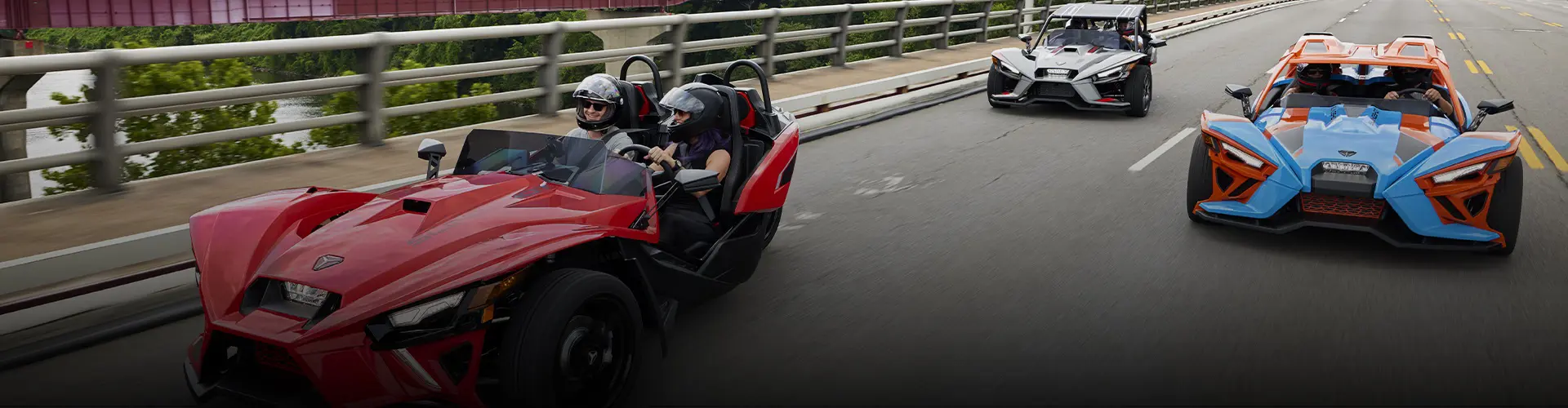 Three colorful Polaris Slingshot autocycles with helmeted riders traveling together across a city bridge.