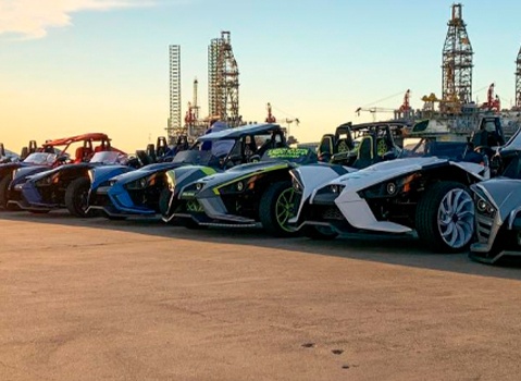 Colorful Polaris Slingshot autocycles lined up near offshore drilling rigs at sunset.