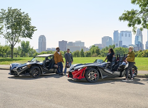 Eight people gather beside four colorful Polaris Slingshot autocycles parked outside an industrial brick building.