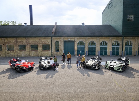 Eight people gather beside four colorful Polaris Slingshot autocycles parked outside an industrial brick building.