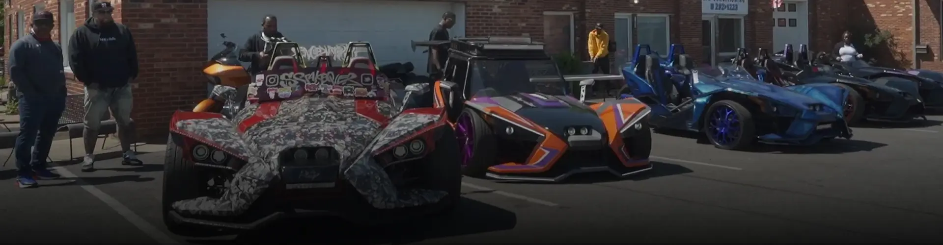 Multiple customized Polaris Slingshot three-wheel vehicles displayed in a parking lot, with attendees gathered nearby.